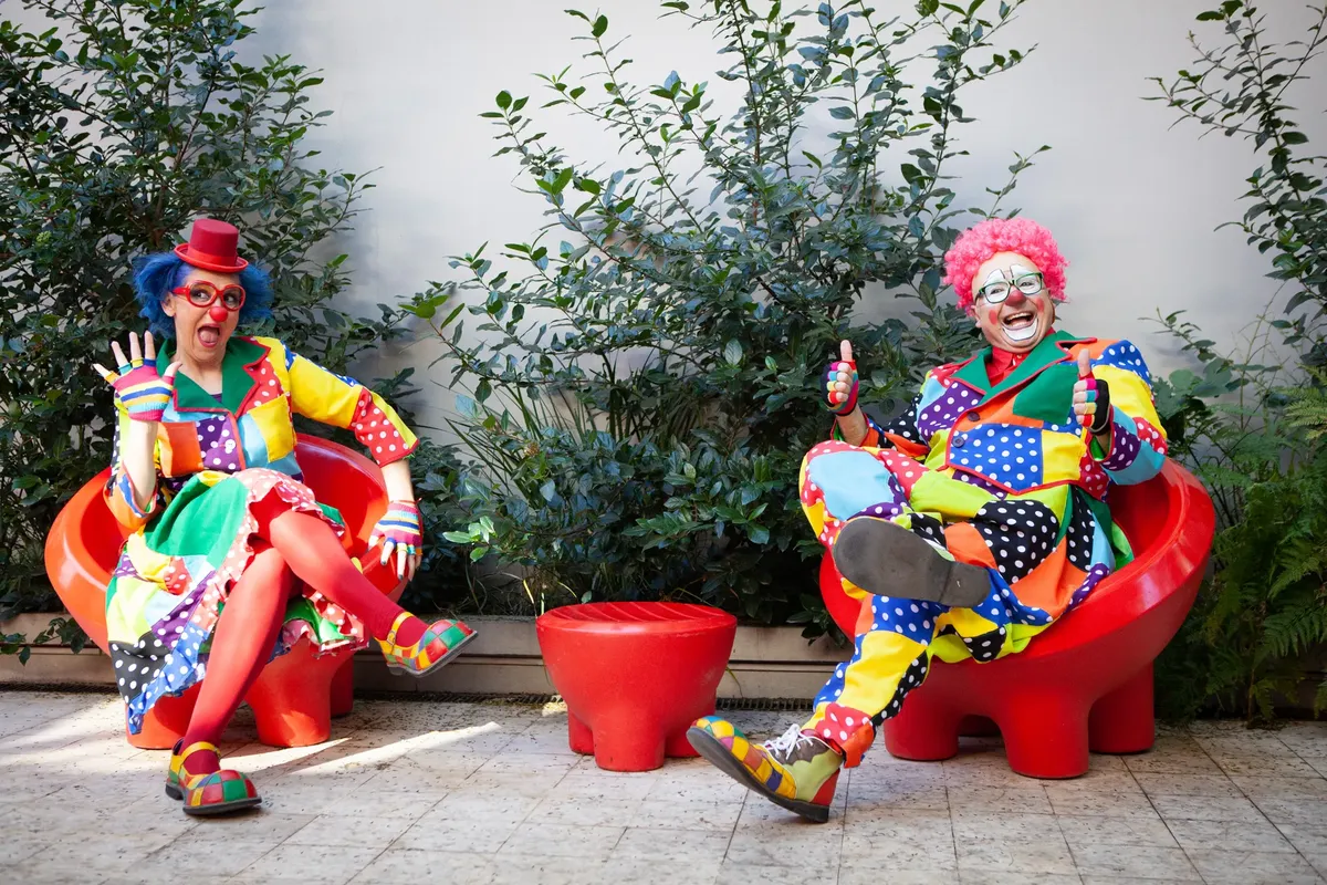 Vibrant clowns in colorful costumes sitting outside with red chairs, spreading joy and humor.