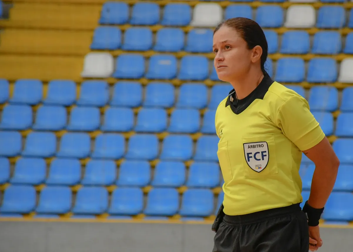 Woman futsal referee in yellow uniform stands focused in empty indoor stadium with blue seats.